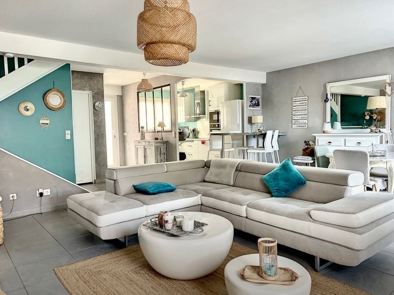 A spacious living area features a modern sectional sofa adorned with teal accent pillows. A round coffee table sits in the center, surrounded by a textured area rug. Light filters in through windows, highlighting the subtle color contrasts of the walls and decor.
