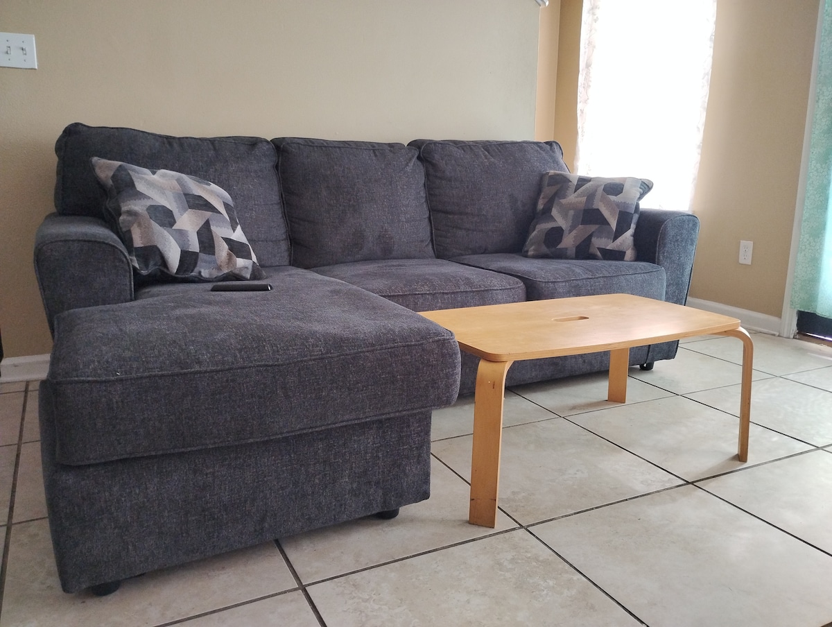 A gray sectional sofa occupies the living area, featuring decorative pillows on either side. In front, a light wood coffee table provides a functional surface. Natural light enters through nearby windows, creating a relaxed setting.