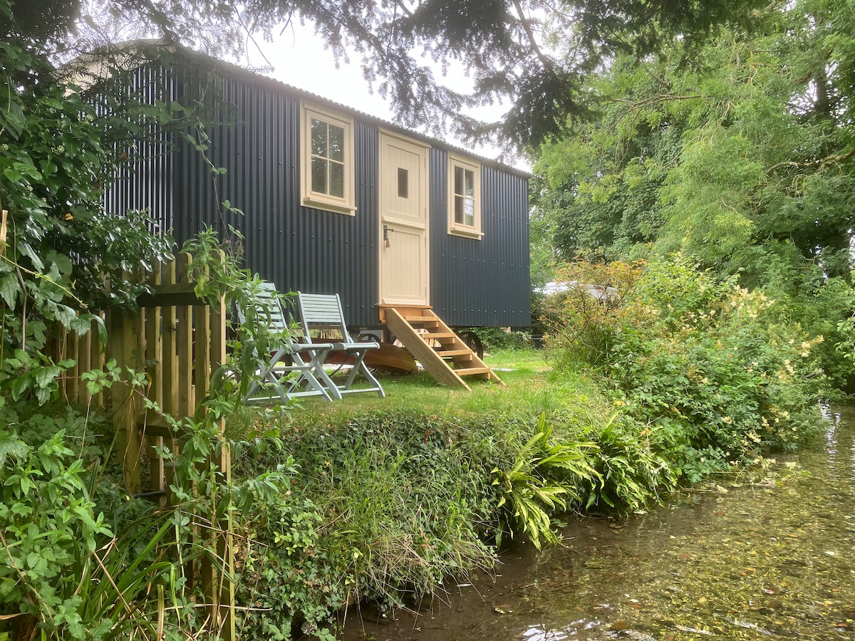 A shepherd's hut is situated amidst lush greenery, framed by trees. A small patio area features two chairs and wooden steps leading to the entrance. A pristine chalk stream flows adjacent to the hut, enhancing the natural setting.