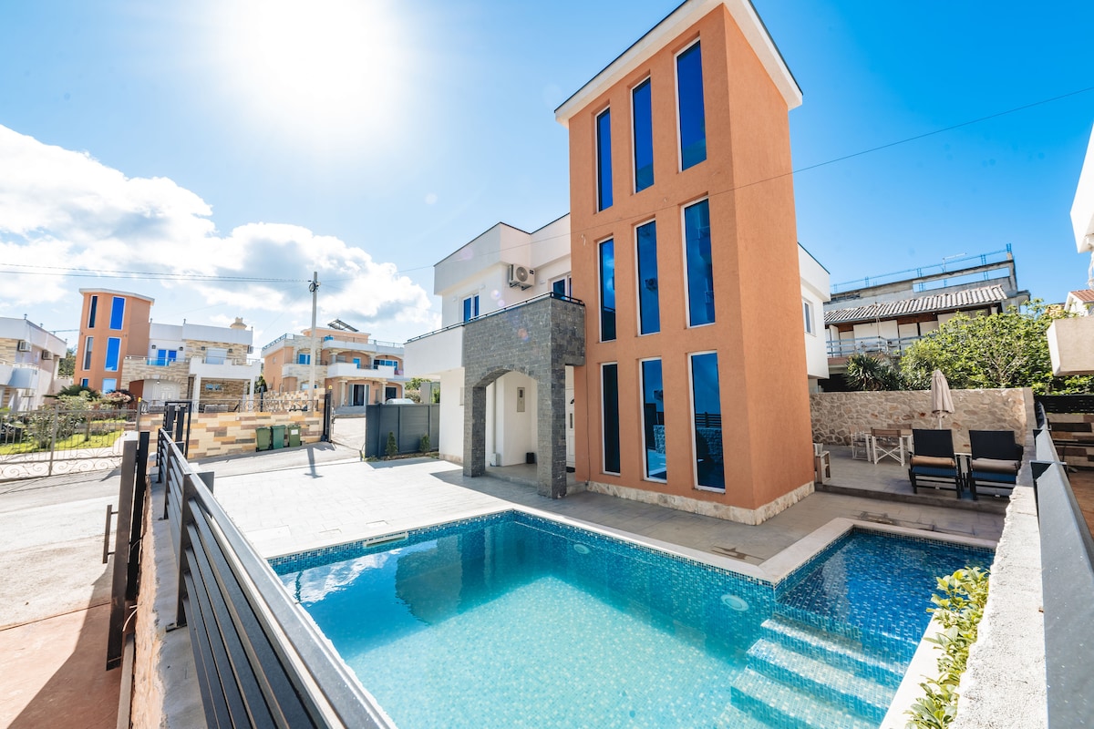 The exterior of a modern building features large glass windows and a striking orange façade. A clear swimming pool reflects the sunlight, bordered by a smooth stone deck and lounge chairs. Surrounding greenery adds to the inviting setting of the space.