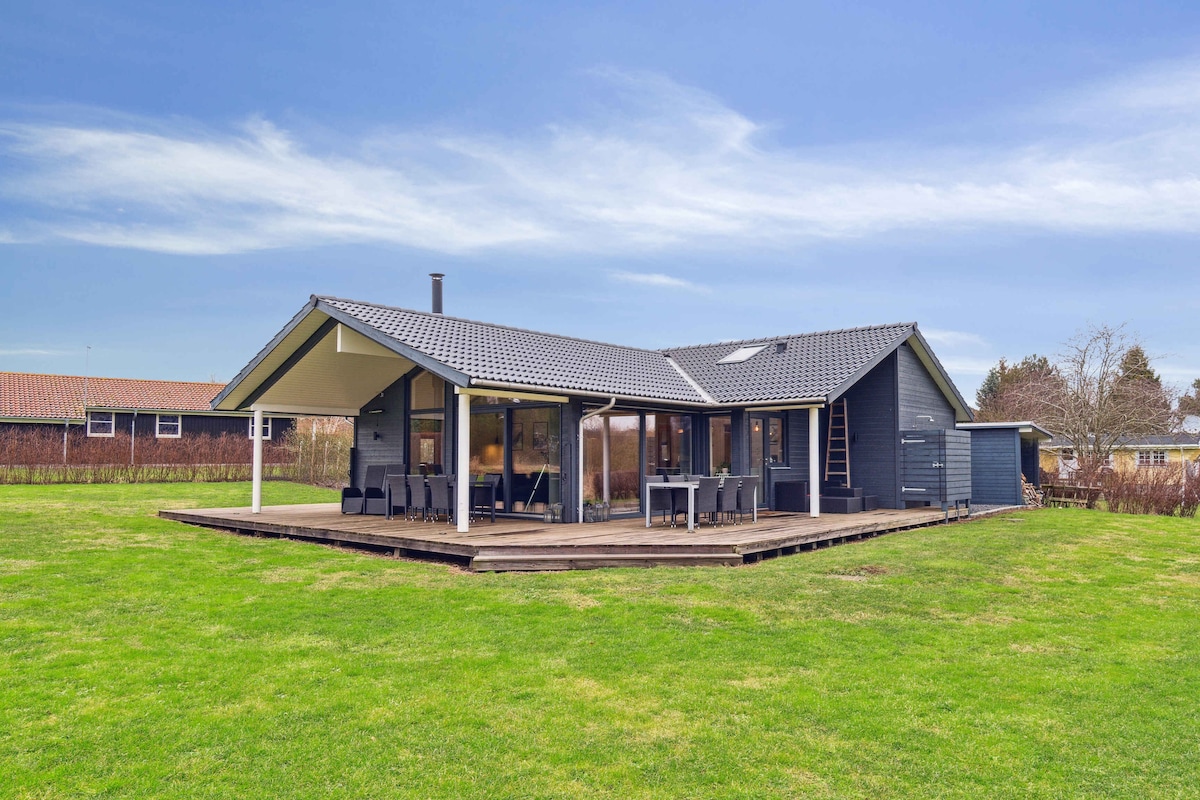 The summer house features a modern design with a spacious, partially covered terrace. A large seating area is visible on the terrace, offering ample space for outdoor gatherings. The building is surrounded by green lawn and includes a chimney and several windows for natural light.