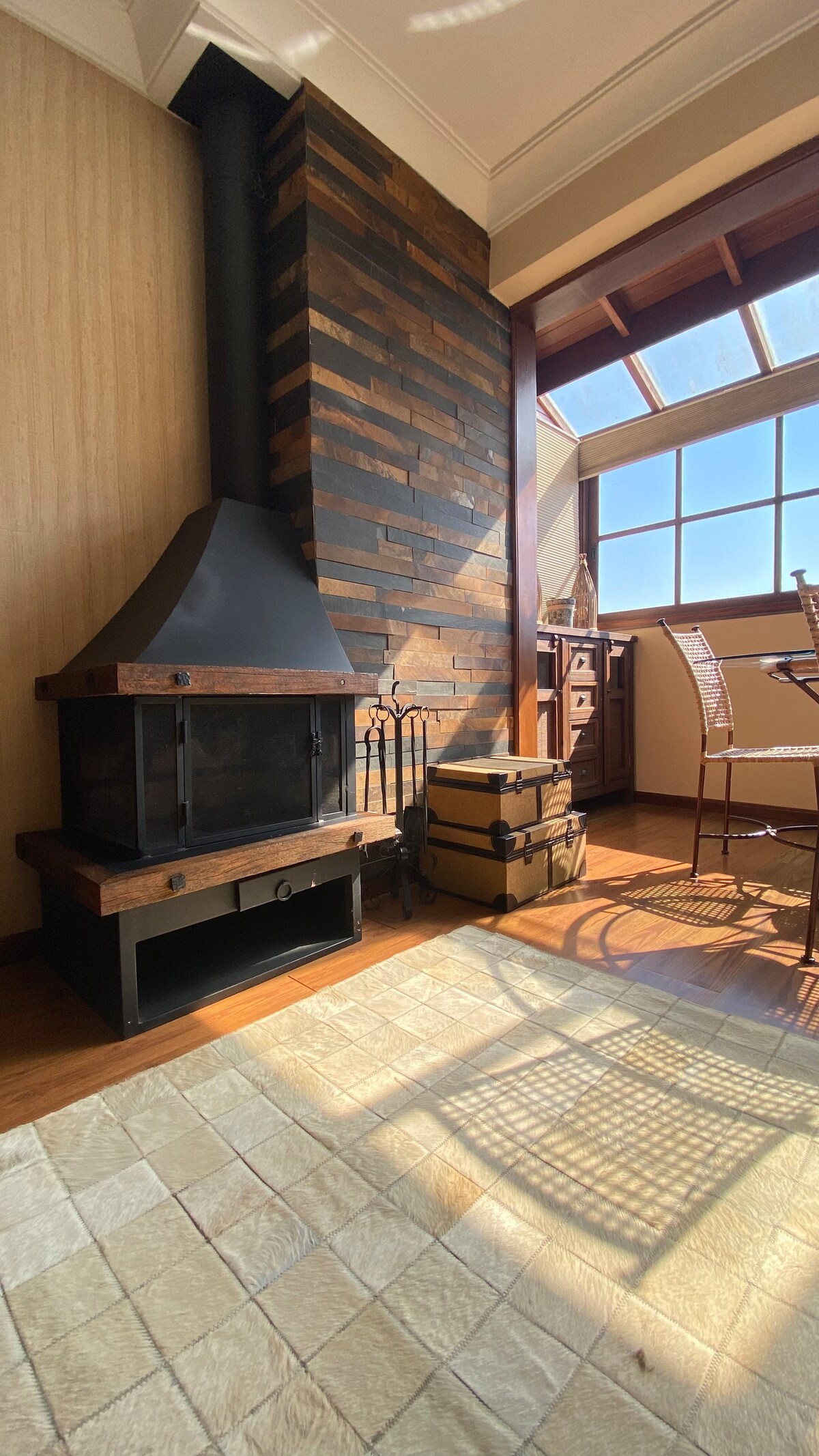 A corner of the living area features a modern fireplace with a dark stone surround, complemented by a wooden floor. Sunlight streams in through large windows, casting soft shadows on the tiled floor. Nearby, woven chairs and decorative storage boxes add character to the space.