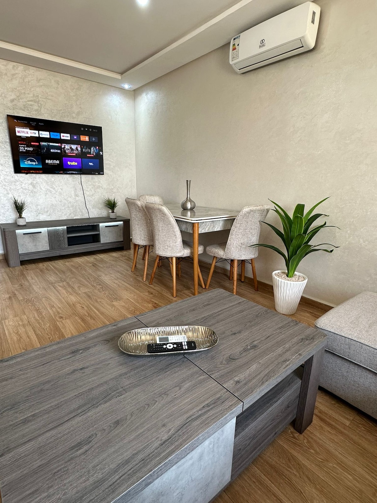 A modern living area features a sleek coffee table in the foreground, complemented by a comfortable sofa nearby. A dining table with four upholstered chairs is positioned under a wall-mounted television, while a decorative plant adds a touch of greenery to the space.