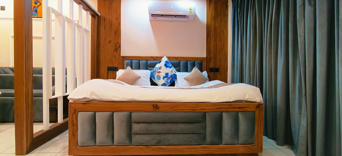 A spacious bed with a wooden frame is positioned against a backdrop of soft-colored walls. Decorative pillows add a touch of style, and air conditioning is positioned above. Floor-to-ceiling curtains frame the area, complementing the overall decor.