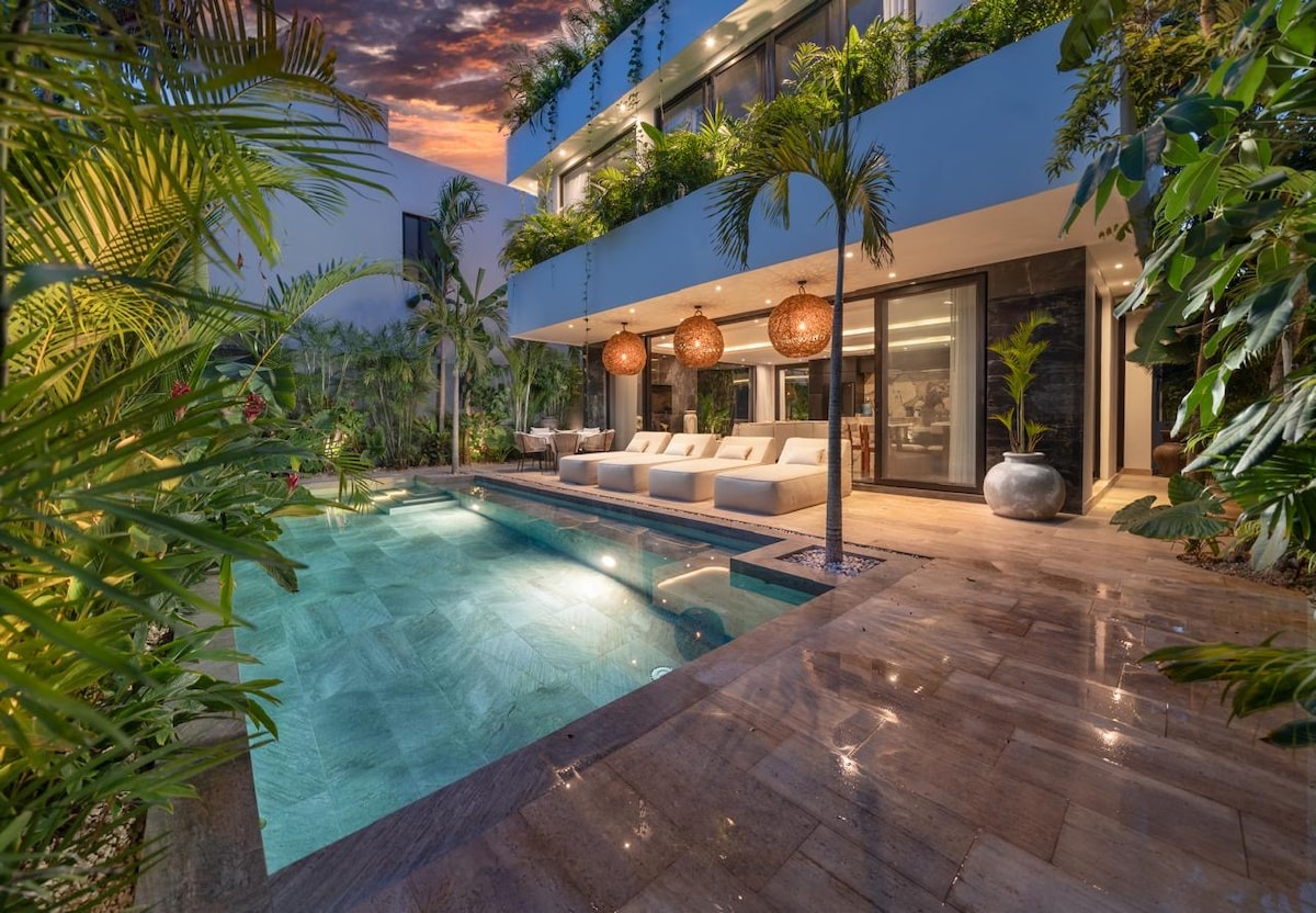 An outdoor area features a tranquil pool framed by lush greenery and elegant furnishings. Sofa loungers provide a relaxing space, while striking pendant lights add a touch of ambiance above. The surrounding plants enhance the serene environment, inviting a sense of relaxation and connection with nature.