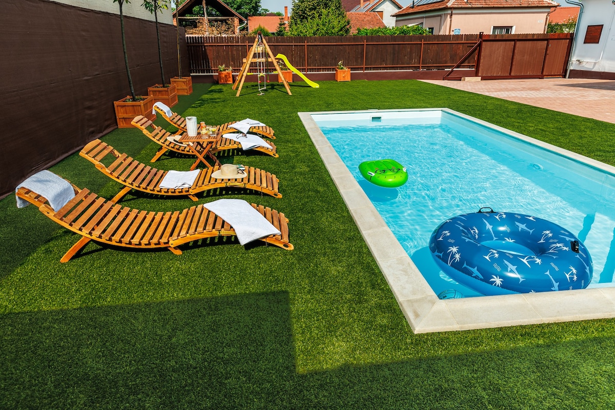 A bright outdoor space is presented with a sparkling pool surrounded by plush green grass. Several wooden lounge chairs are arranged for relaxing, along with a yellow slide in the background. Two inflatable rings float on the water, creating a welcoming aquatic setting.