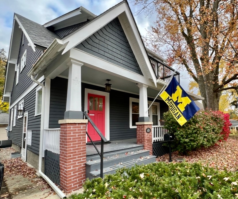 The exterior of a historic home is depicted, featuring a covered porch with a bright pink door. A flag representing the University of Michigan is displayed prominently. Colorful autumn leaves and well-maintained landscaping enhance the welcoming appearance of the property.