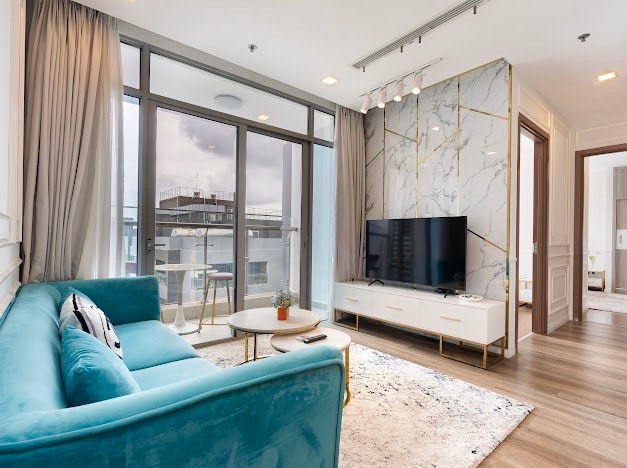 A modern living area features a blue sofa complemented by a round coffee table. A large television is mounted on a stylish cabinet against a marble accent wall. Floor-to-ceiling windows allow natural light and provide views of the outdoors.