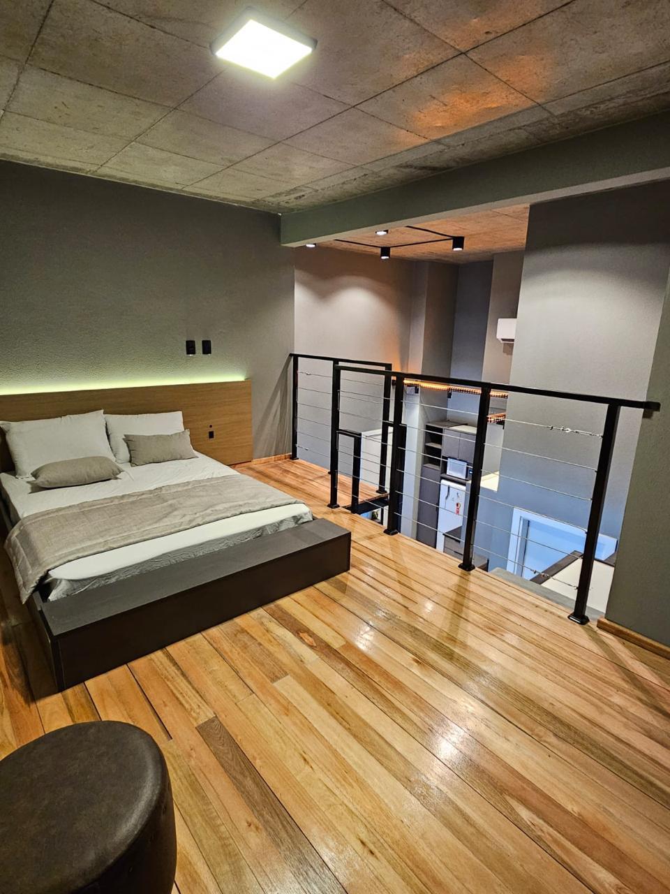 A loft bedroom is elevated above a spacious living area, featuring a large bed with neutral bedding. Smooth wooden flooring contrasts with concrete walls, while a minimalist railing provides safety and defines the space. Soft lighting enhances the room's modern design.
