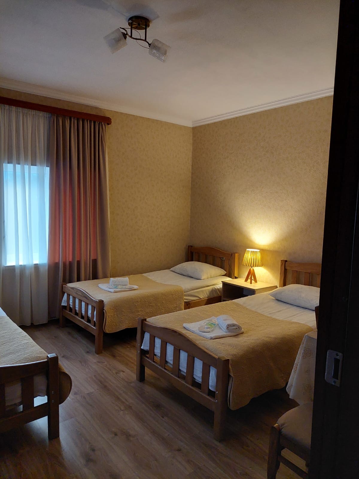 A well-lit room contains three wooden beds, each topped with neatly arranged yellow blankets. Soft pillows are placed on the beds. A small lamp provides warm light, while a window with sheer curtains allows natural light to fill the space, enhancing the cozy ambiance.