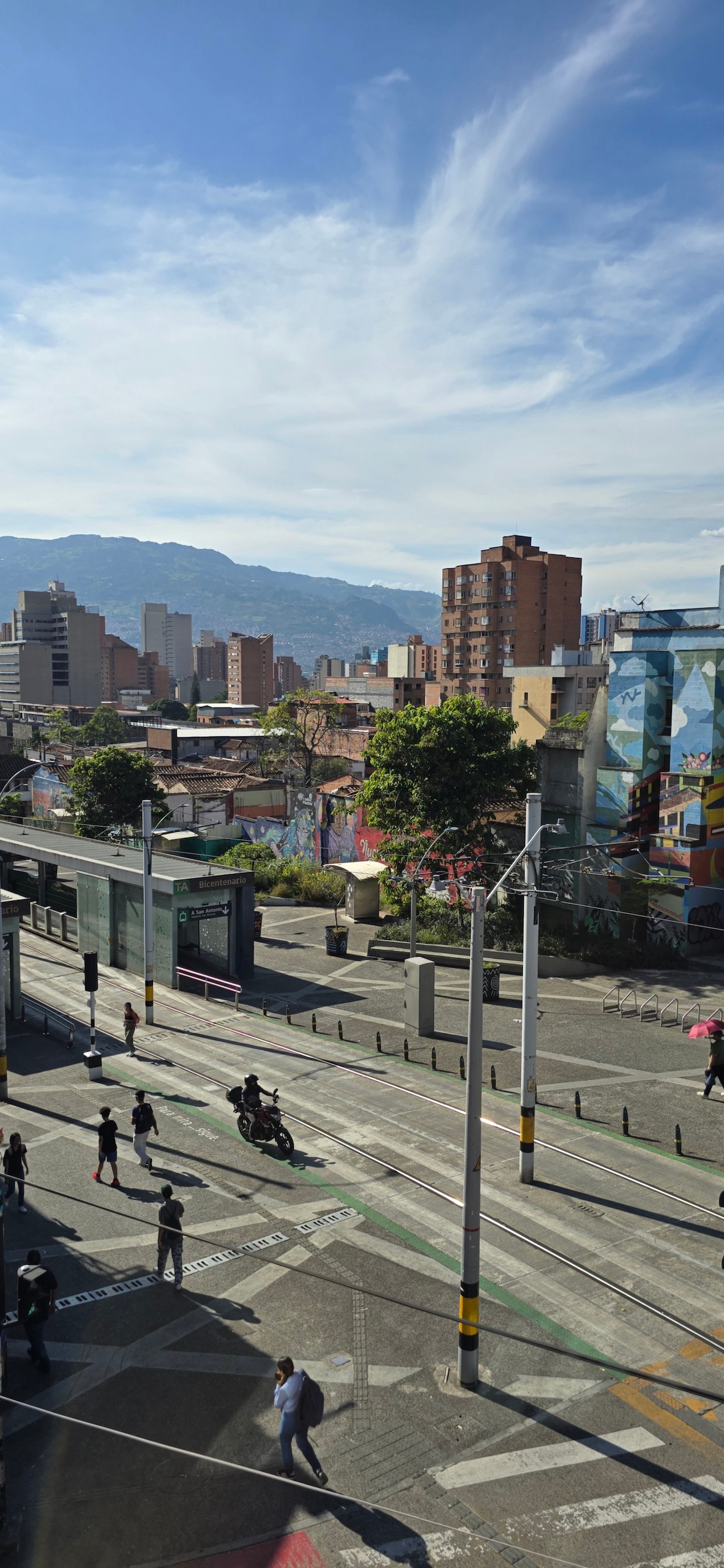 A vibrant urban scene is captured, featuring a lively street populated with pedestrians and cyclists. The backdrop includes a mix of contemporary buildings and colorful murals, while distant mountains stand against a clear blue sky, highlighting the dynamic city landscape.