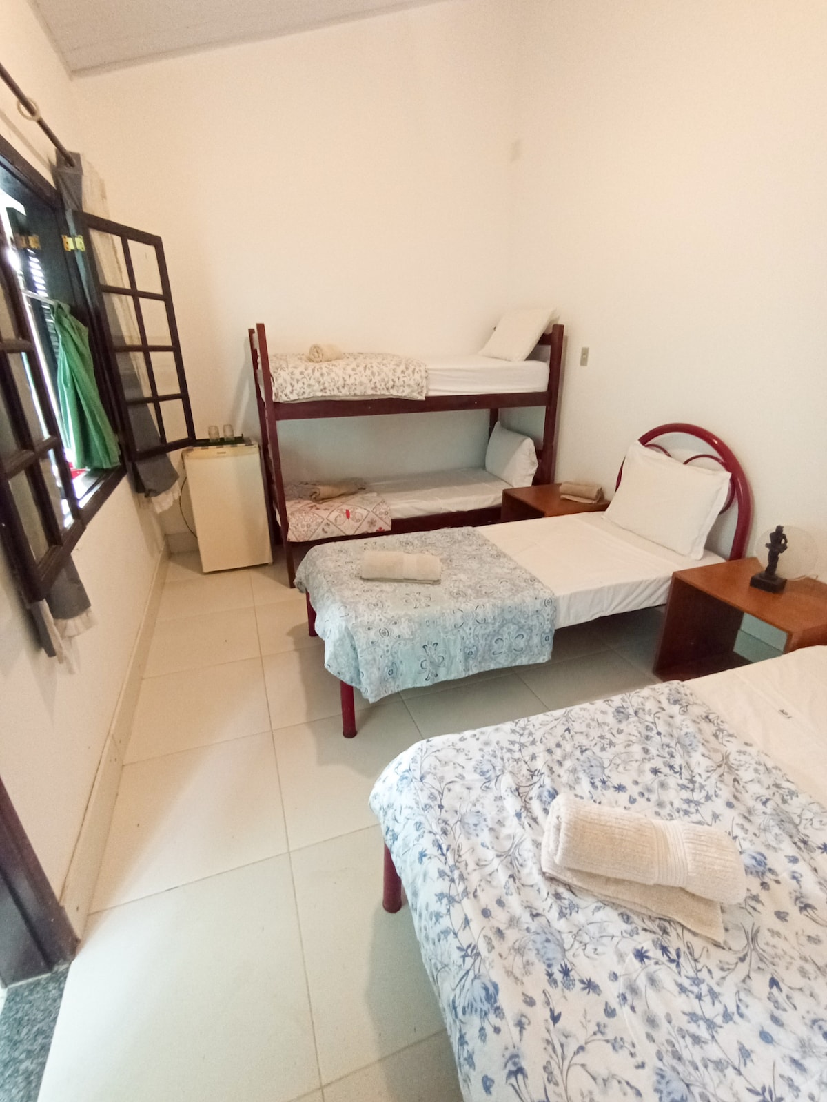 A bright room features a bunk bed with white bedding and a single spacious bed adorned with a floral blanket. A bedside table is positioned next to the single bed, and soft towels are neatly arranged. Light enters through a window with wooden shutters, enhancing the airy feel.