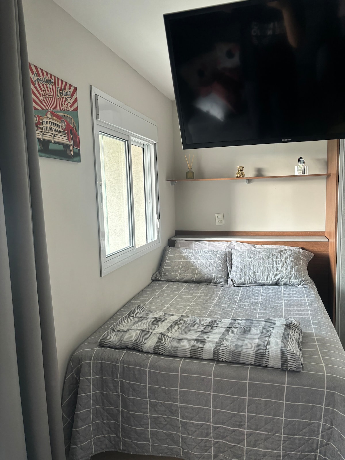 A compact sleeping area features a spacious bed covered with a textured gray blanket. A television is mounted above, and natural light filters through a nearby window, which is flanked by curtains. A small shelf holds decorative items, enhancing the room's functionality.