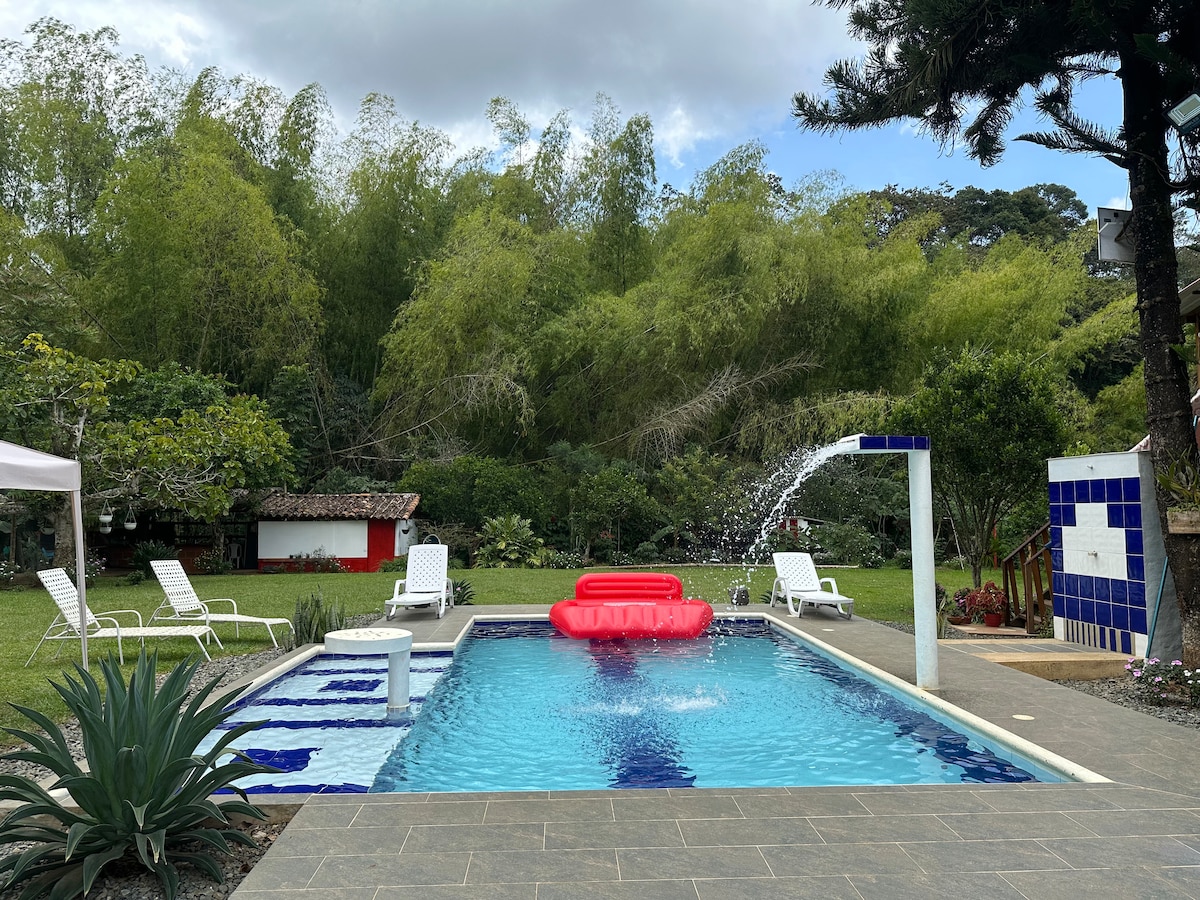 A sparkling pool is surrounded by a lush garden, featuring a vibrant red float and white lounge chairs. A fountain adds a refreshing touch, while various trees and shrubs provide natural beauty in the outdoor space.