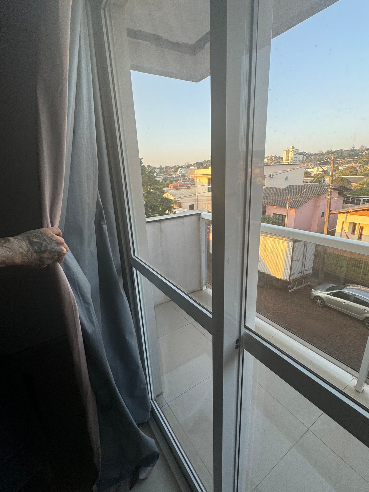 A view from a window shows a city landscape featuring a mix of residential buildings and vehicles. Natural light filters through sheer curtains, illuminating the interior. The balcony space is accessible, with a concrete floor visible outside.