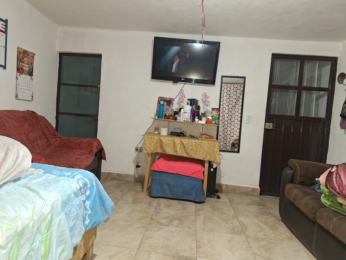 A living area is presented with two sofas, one covered in colorful blankets. A television is mounted on the wall, and a small table holds personal items. Two doors are visible, one leading outside and the other into a different room. The flooring is tiled.