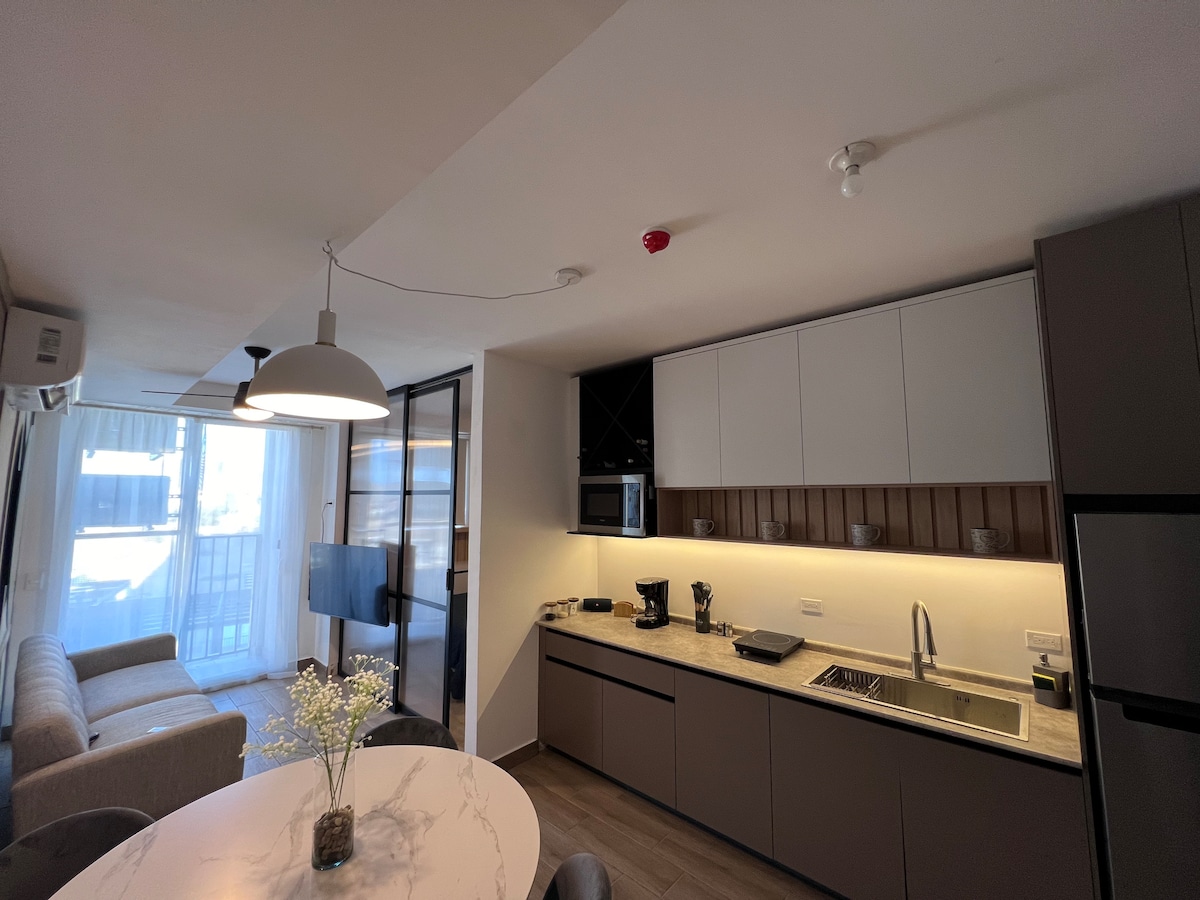 A modern kitchen area is featured, equipped with sleek cabinetry and essential appliances, including a coffee maker. A dining table is positioned nearby, with fresh flowers as a centerpiece. Natural light filters in through a large window, illuminating the connected living space.