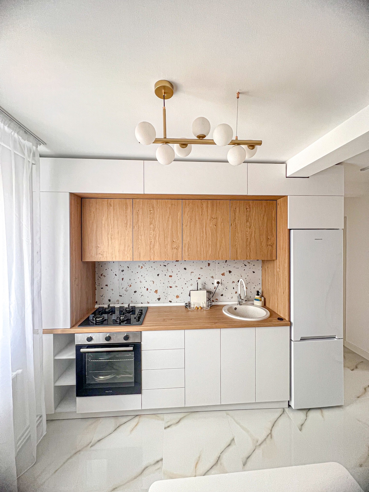 A modern kitchen features sleek cabinetry in white and oak, complemented by a decorative backsplash with colorful specks. Essential appliances, including a stove and refrigerator, are integrated into the design. Ample counter space is available for meal prep, and natural light enters through sheer curtains.