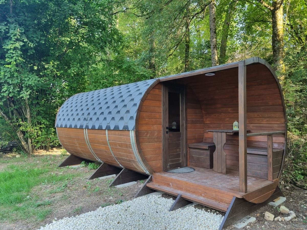 A wooden barrel-shaped structure is nestled among lush greenery. The entrance features a simple wooden door and a small covered patio with a table and benches. Surrounding the cabin, a natural landscape adds to the serene environment.