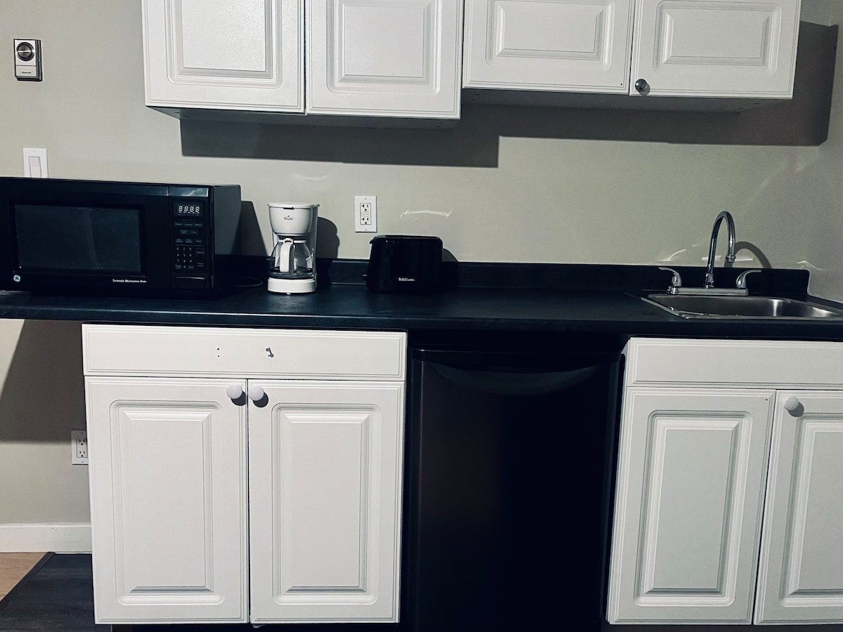 A compact kitchen area features white cabinetry and a dark countertop. Appliances include a microwave, coffee maker, and toaster, alongside a sink and refrigerator. The layout provides functional space for meal preparation, with essential dish-wares and glasses available.