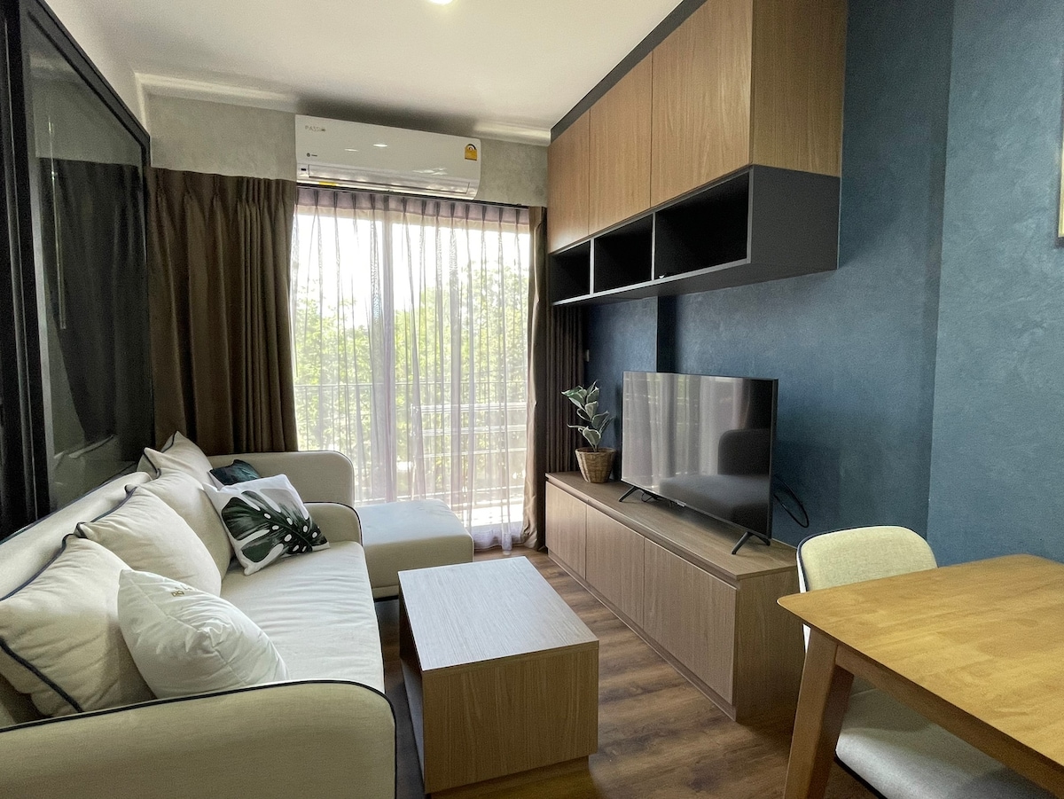 A living area is presented with a light-colored sofa, a coffee table, and a contemporary television mounted on a wooden unit. Sunlight filters through sheer curtains, illuminating the space. A potted plant adds a touch of greenery, complementing the warm wood flooring.