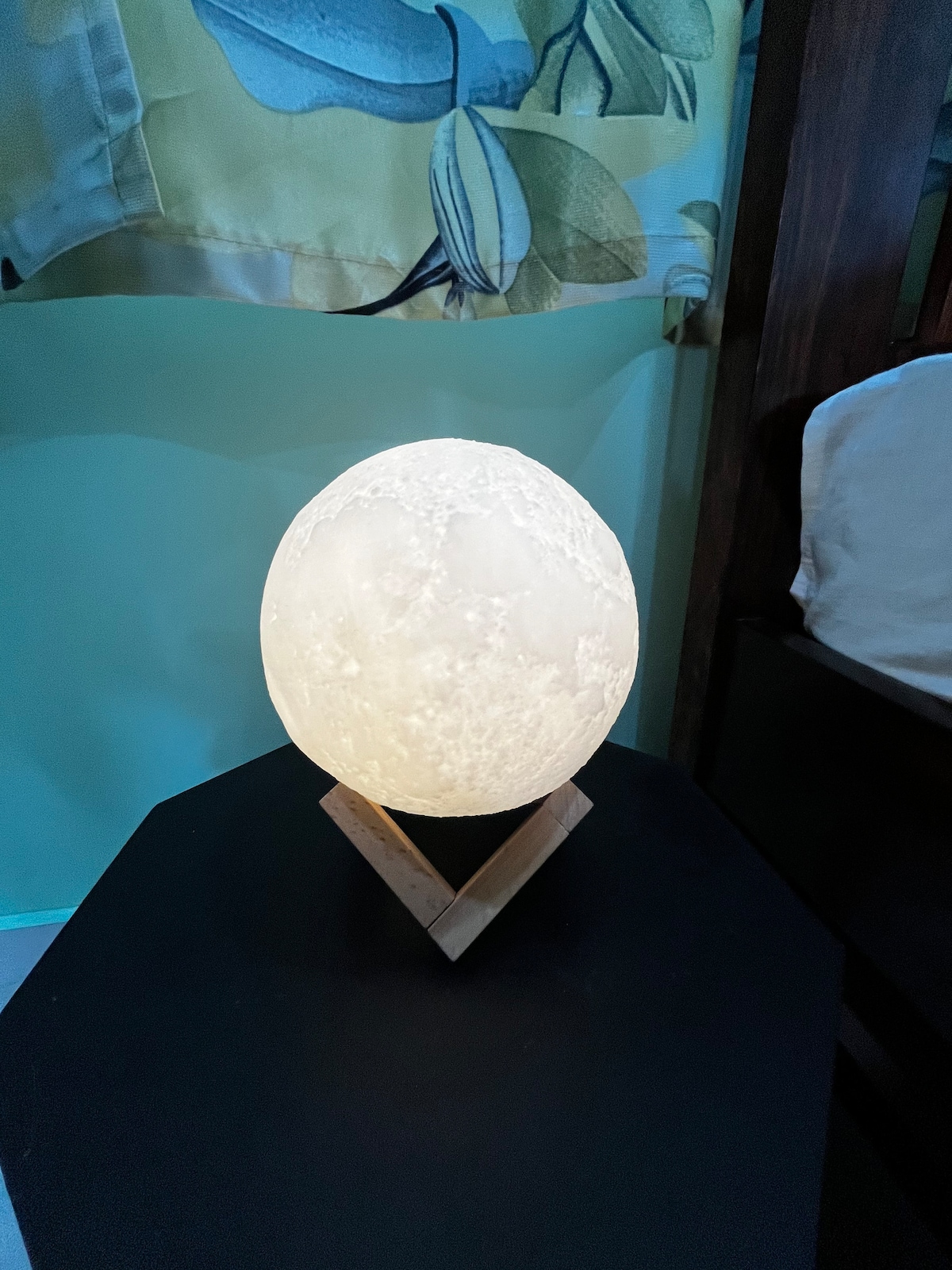 A unique lamp in the shape of an egg emits a soft, warm glow, creating a calming ambiance. It is positioned on a simple triangular wooden stand, contrasting with the surrounding decor, which features light-colored curtains decorated with leaf patterns.
