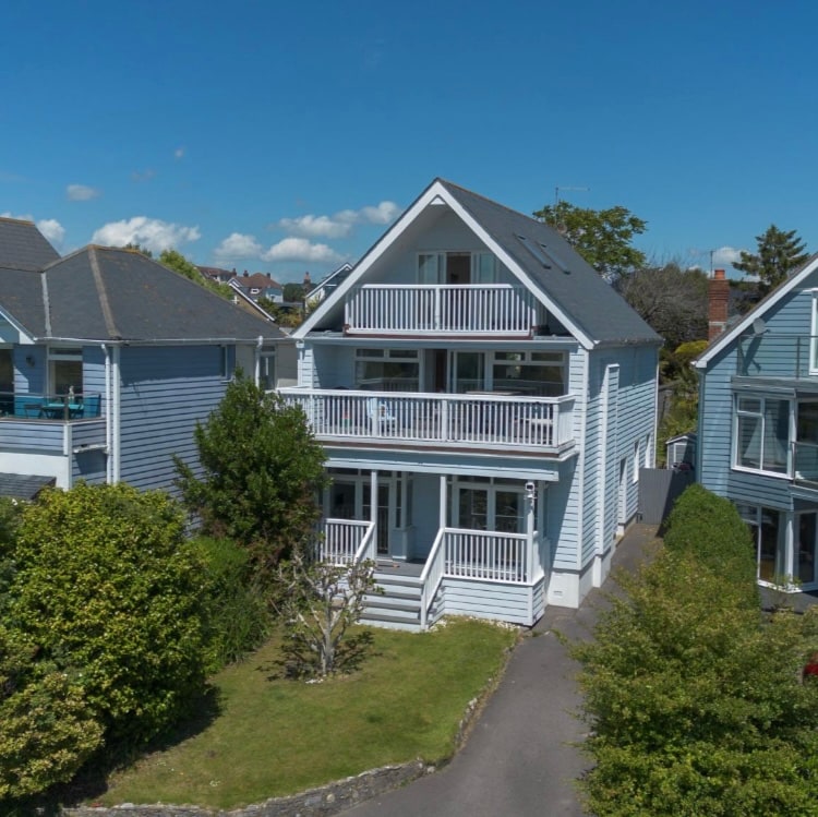 A three-story New England Style home with a light blue exterior is surrounded by greenery. The house features multiple balconies on the upper levels and a welcoming front porch. The driveway leads to a well-maintained garden area, enhancing the overall appeal of the property.