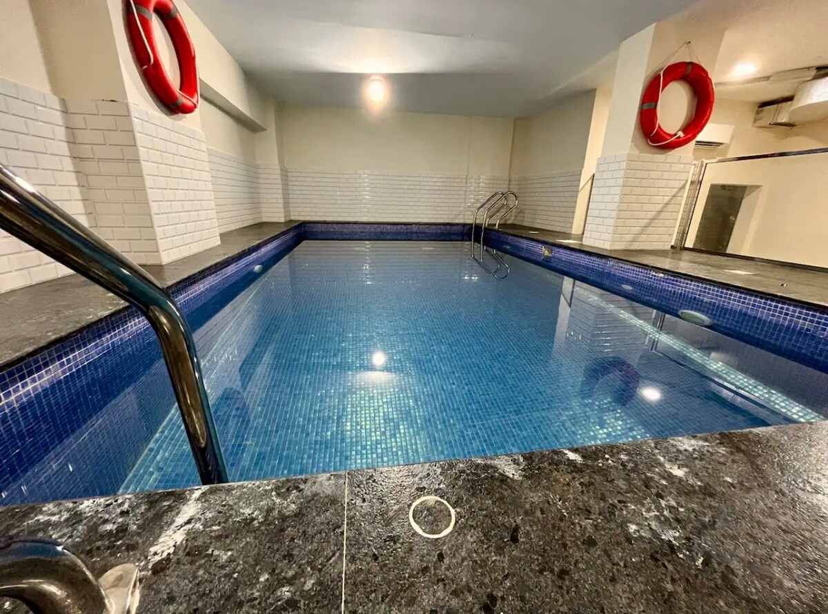 An indoor swimming pool is surrounded by sleek gray tiling and features bright blue mosaic tiles on the pool walls. Life rings are affixed to the walls for safety. The lighting illuminates the water, creating a tranquil ambiance.