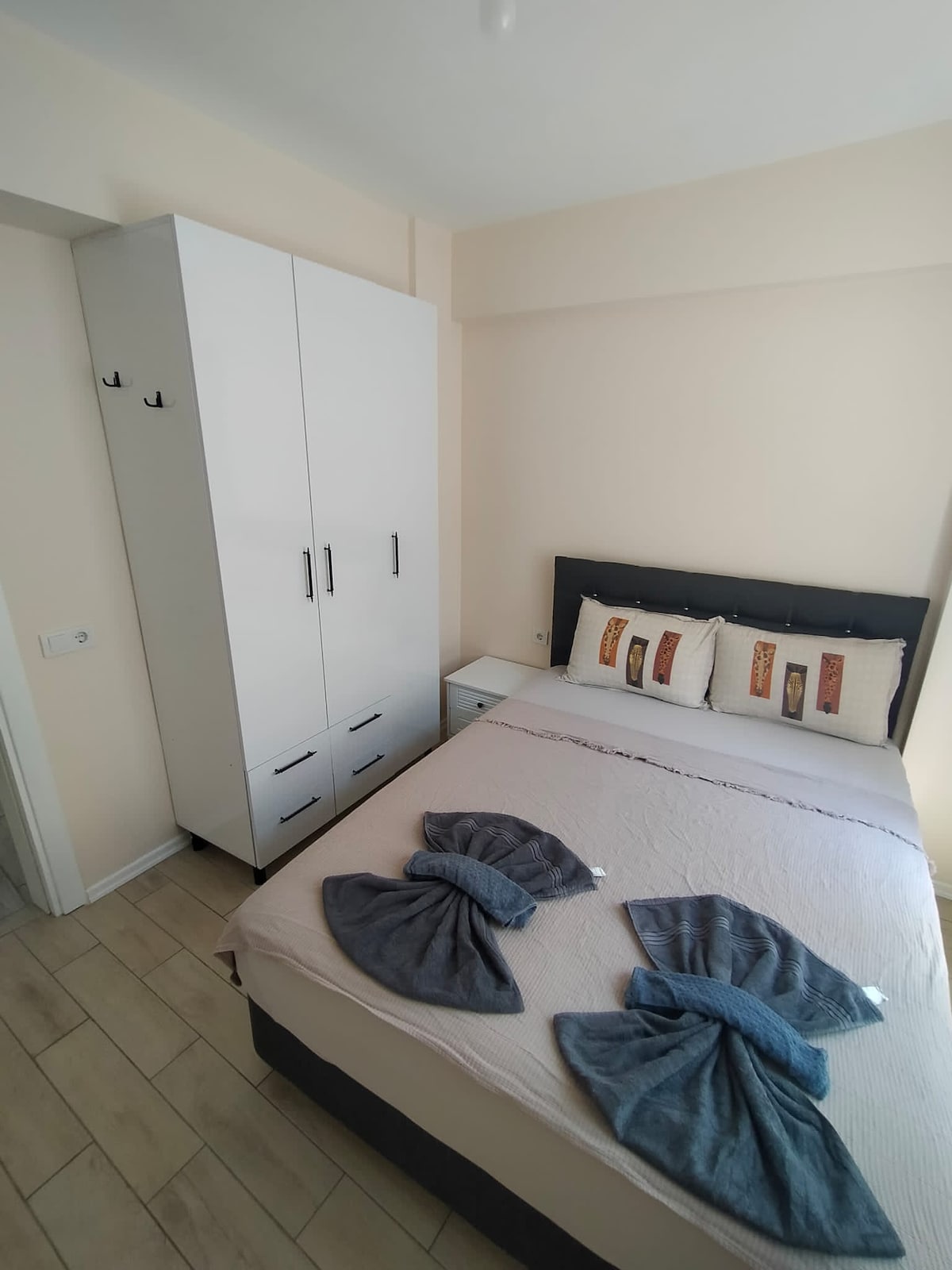 A modest bedroom features a spacious bed adorned with neatly arranged gray towels. A light-colored wardrobe stands against the wall, complemented by a small nightstand. Decorative pillows with subtle patterns add a touch of character to the bed, illuminated by natural light from the nearby window.