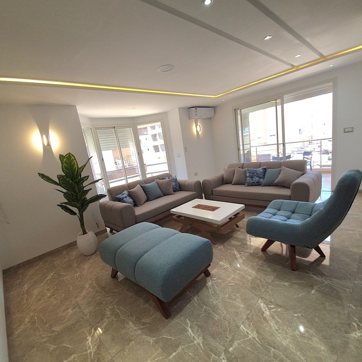 A spacious living area features light marble flooring and modern furniture. A large, neutral-toned sofa is complemented by an accent chair and an ottoman. Natural light floods the room through large windows, enhancing a sense of openness. A plant in a decorative pot adds a touch of greenery.