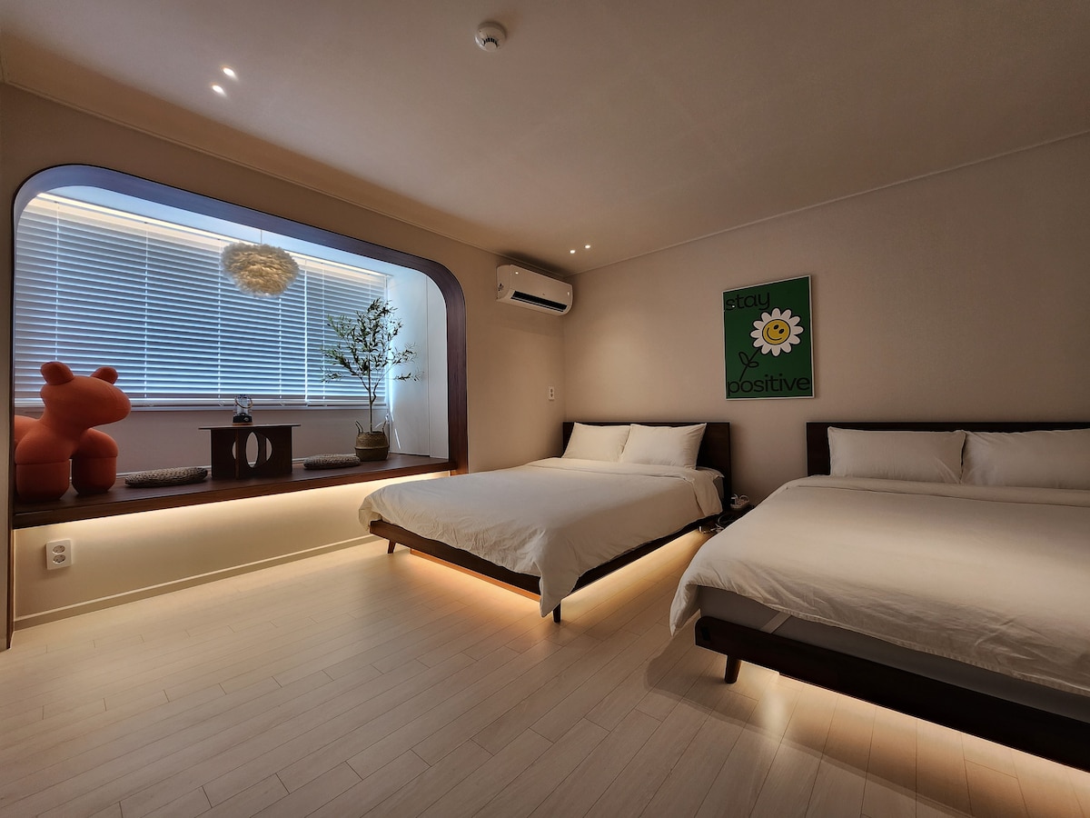 A spacious room features two beds with soft, white bedding. Natural light enters through large windows, complemented by a decorative plant. A comfortable seating area and artwork adorn the space, which is accented by soft lighting along the floor, creating a calm ambiance.