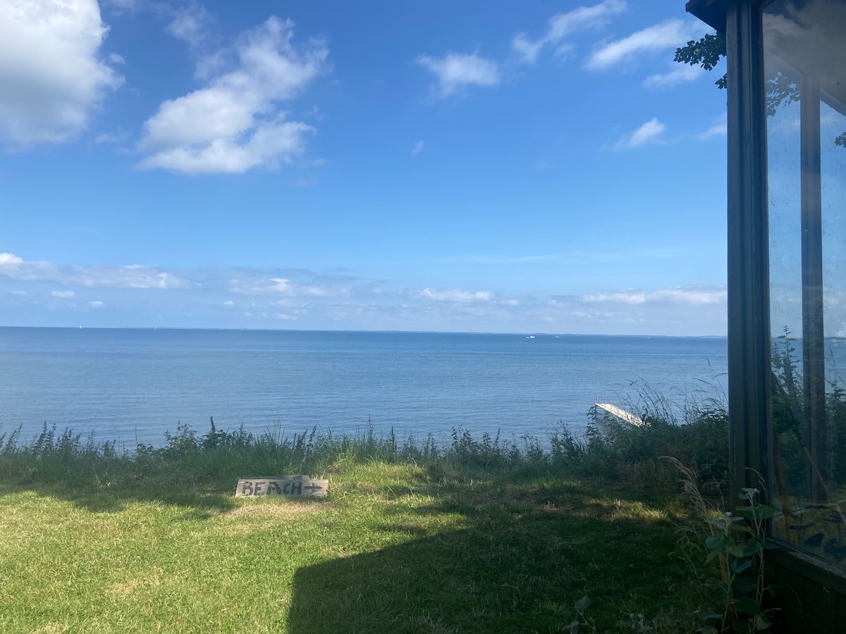 A serene view of the Isefjord is presented, showcasing calm waters under a blue sky with scattered clouds. Lush green grass leads down to the water's edge, with a sign indicating 'BEACH' visible among the natural landscape.