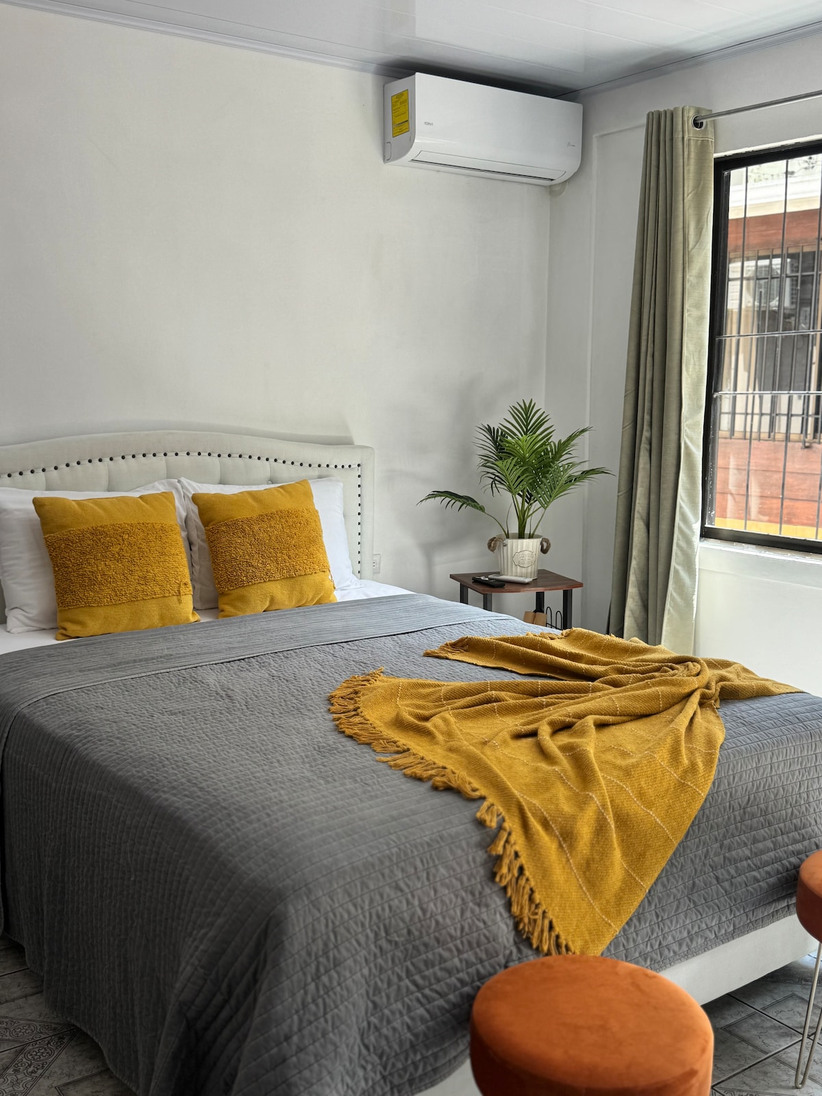 A comfortable bedroom is featured, showcasing a spacious bed adorned with textured throw pillows and a soft blanket. A small table holds a potted plant, while bright curtains frame the window that provides natural light. Two orange stools are positioned nearby.