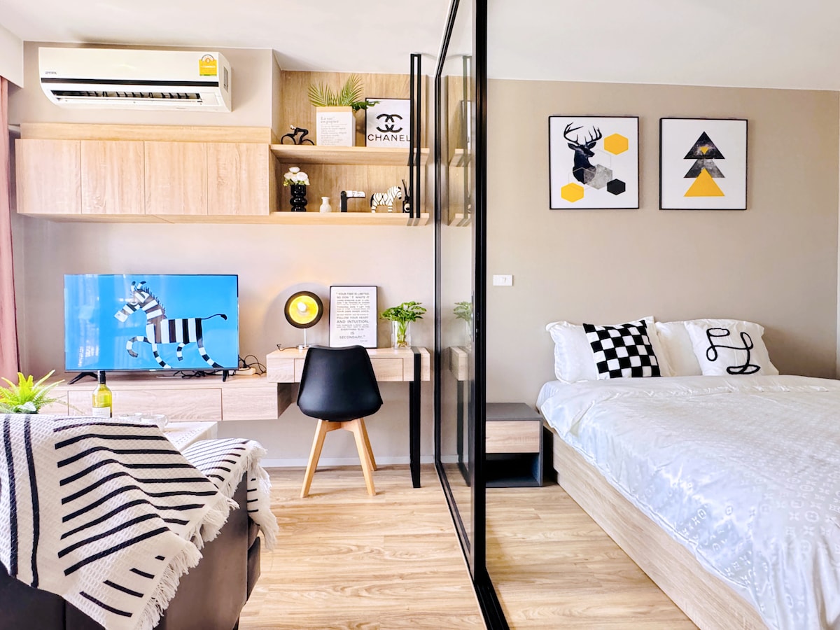 The open studio layout features a spacious bed and a workstation with a black chair. A television is positioned against a light wood accent wall. Decorative elements include framed art and plants, creating a stylish atmosphere complemented by ample natural light.