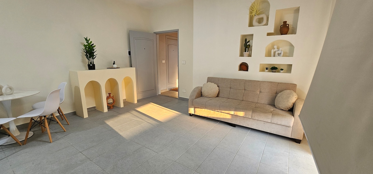A light-filled living area features a beige sofa with decorative cushions, complemented by a round dining table and white chairs. Shelves on the wall display curated decor items. The neutral tones and open layout contribute to a spacious atmosphere.
