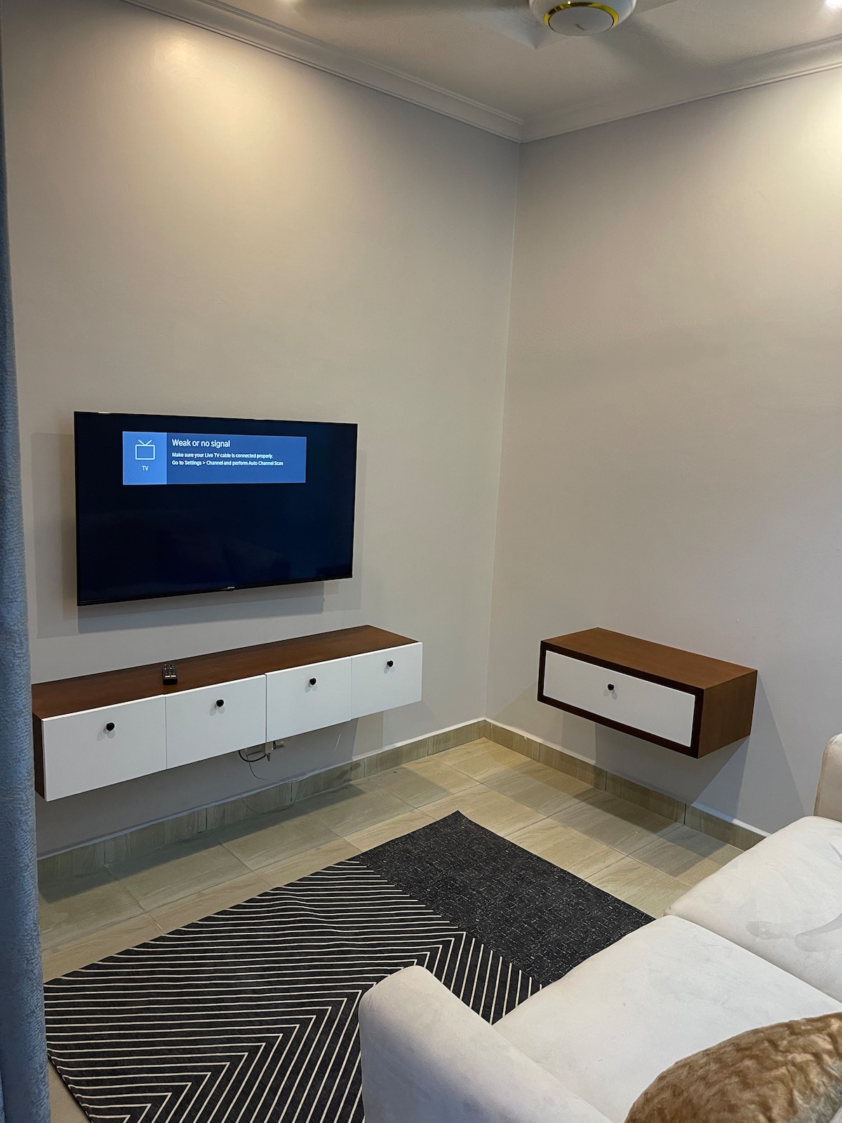 A clean and modern sitting area features a wall-mounted television above a contemporary media console. A small floating shelf is positioned beside the console. A textured area rug adds warmth to the space, complementing the light-colored walls and furniture.