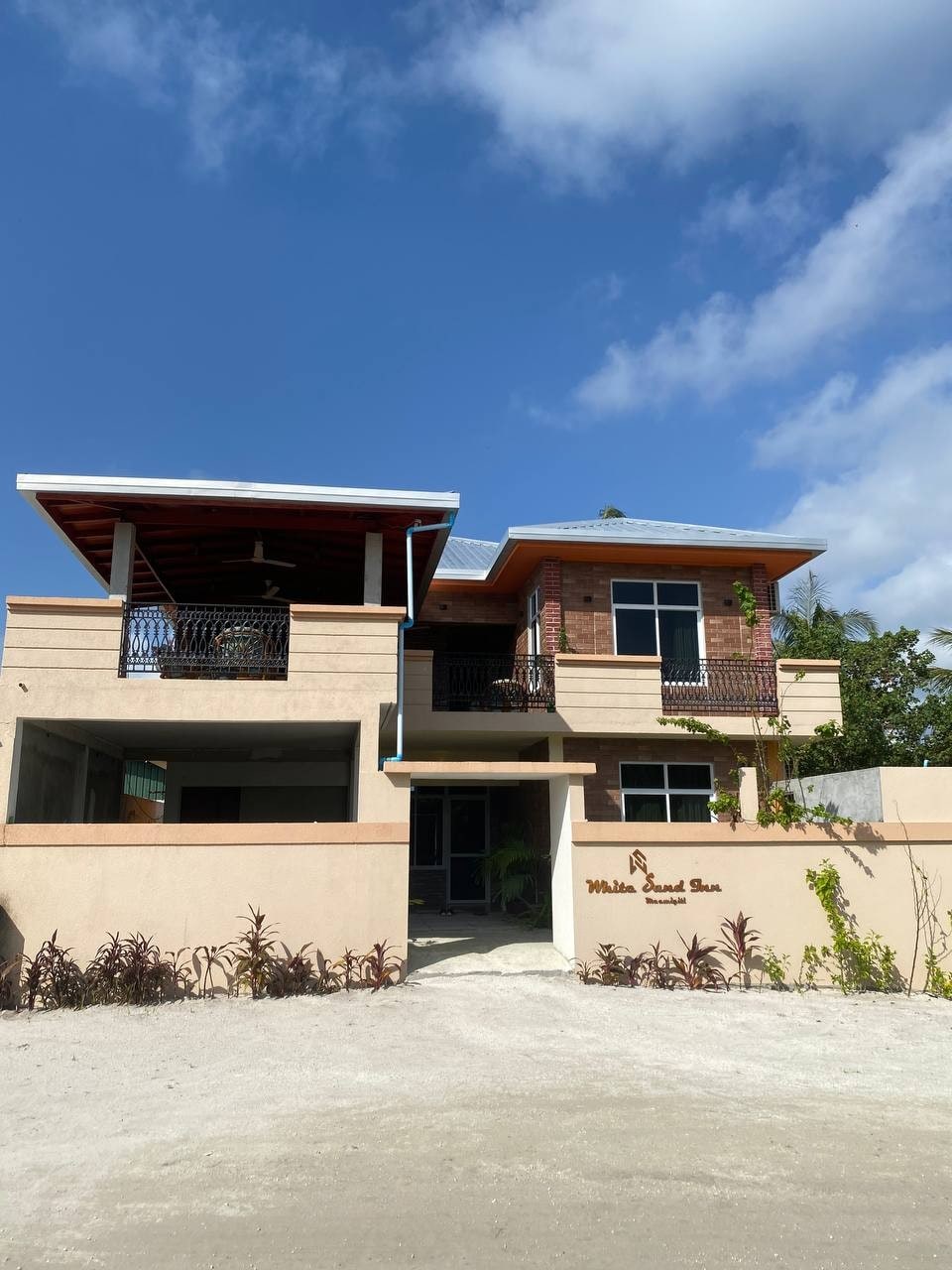 The exterior of a two-story building is showcased, featuring a combination of brick and stucco walls. A covered balcony with decorative railings is visible on the upper level, and a spacious entryway is highlighted below. Lush greenery surrounds the property, enhancing the welcoming presence.