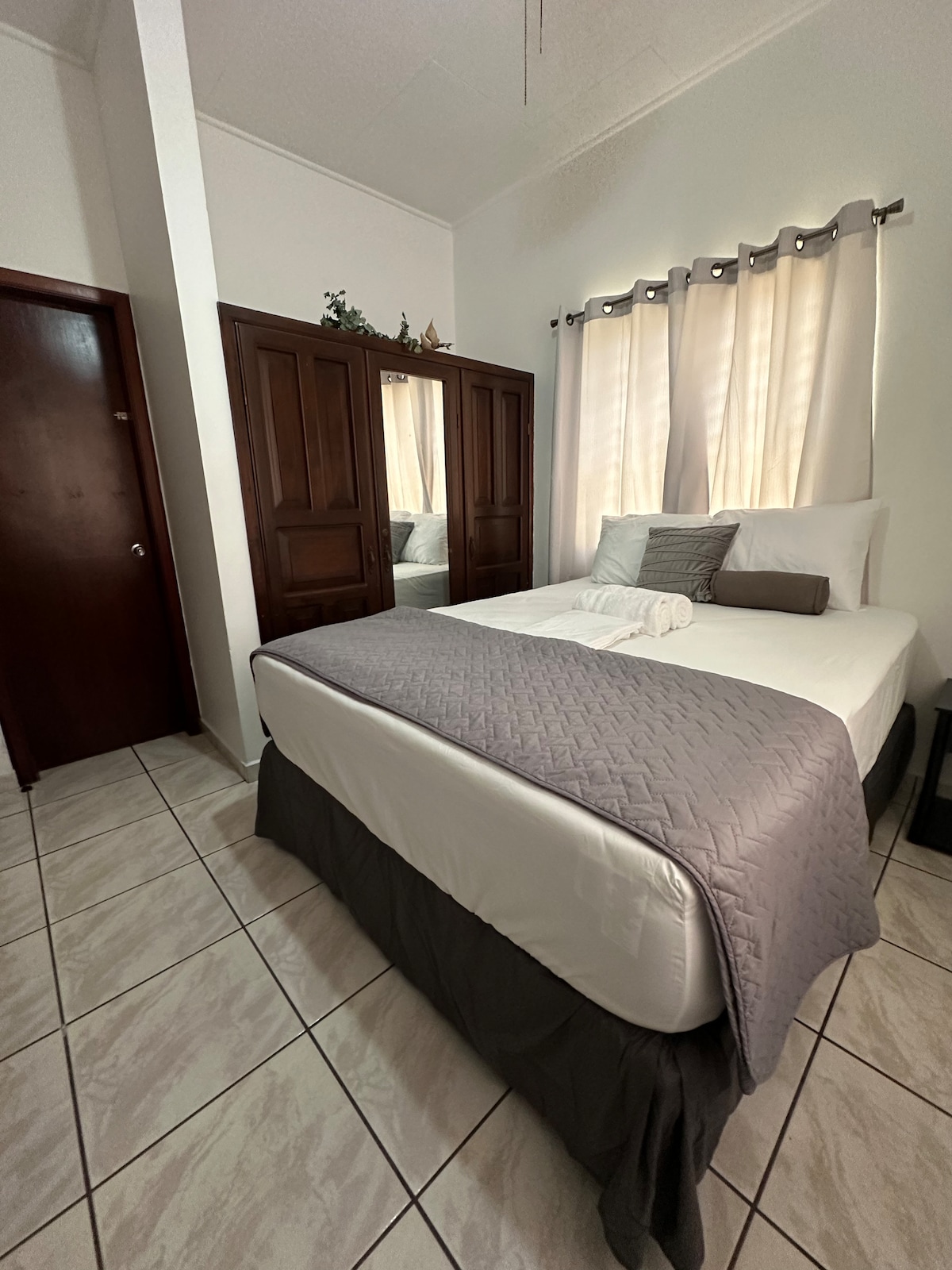 A cozy bedroom features a spacious bed with neutral-colored bedding and decorative pillows. Light-colored curtains frame a window, allowing natural light to fill the room. A mirrored wardrobe is positioned against the wall, enhancing the sense of space with its reflective surface.