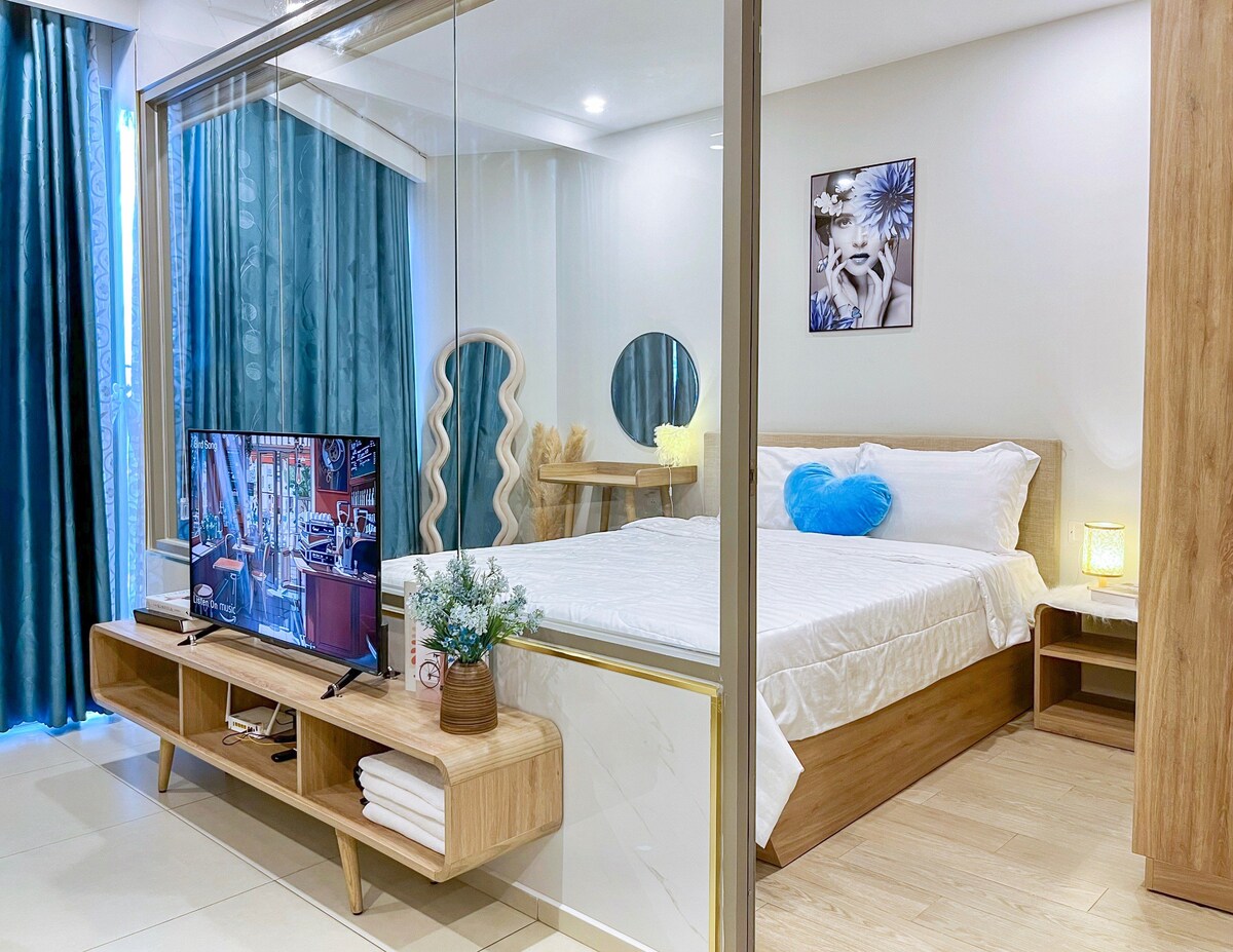 A serene bedroom area is visually separated by glass panels, featuring a spacious bed with white linens and a blue heart-shaped pillow. A wooden media console holds a television, while a round mirror reflects the space. Soft curtains frame the windows, enhancing natural light.