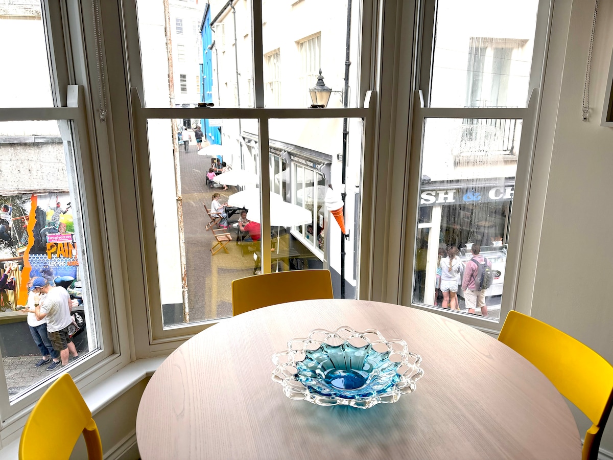 Natural light fills the dining area, where a round table is set with a decorative blue glass bowl. Large windows offer views of the lively street below, featuring café seating and pedestrians enjoying the vibrant atmosphere of the town.