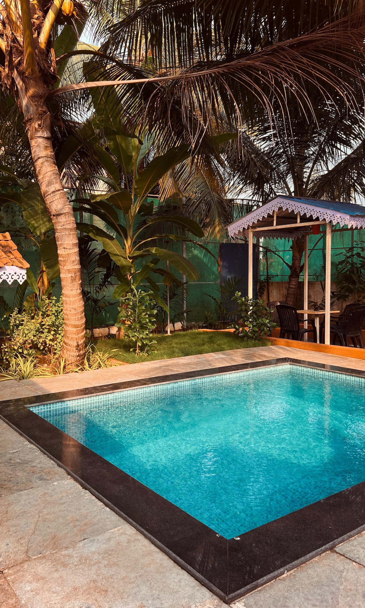 A refreshing pool is surrounded by lush greenery and tall palm trees. A charming pavilion with seating is situated nearby, offering a serene spot for relaxation. The area is designed with stone tiles, and the clear water reflects the sunlight beautifully.