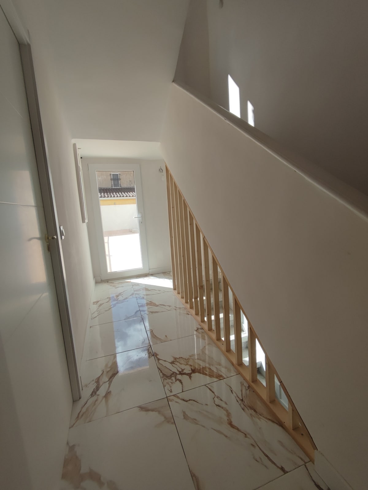 A well-lit stairway features a minimalist design with white walls and a wooden railing. Large windows allow natural light to fill the space, illuminating the glossy marble flooring. A glass door at the end provides access to an outdoor area.