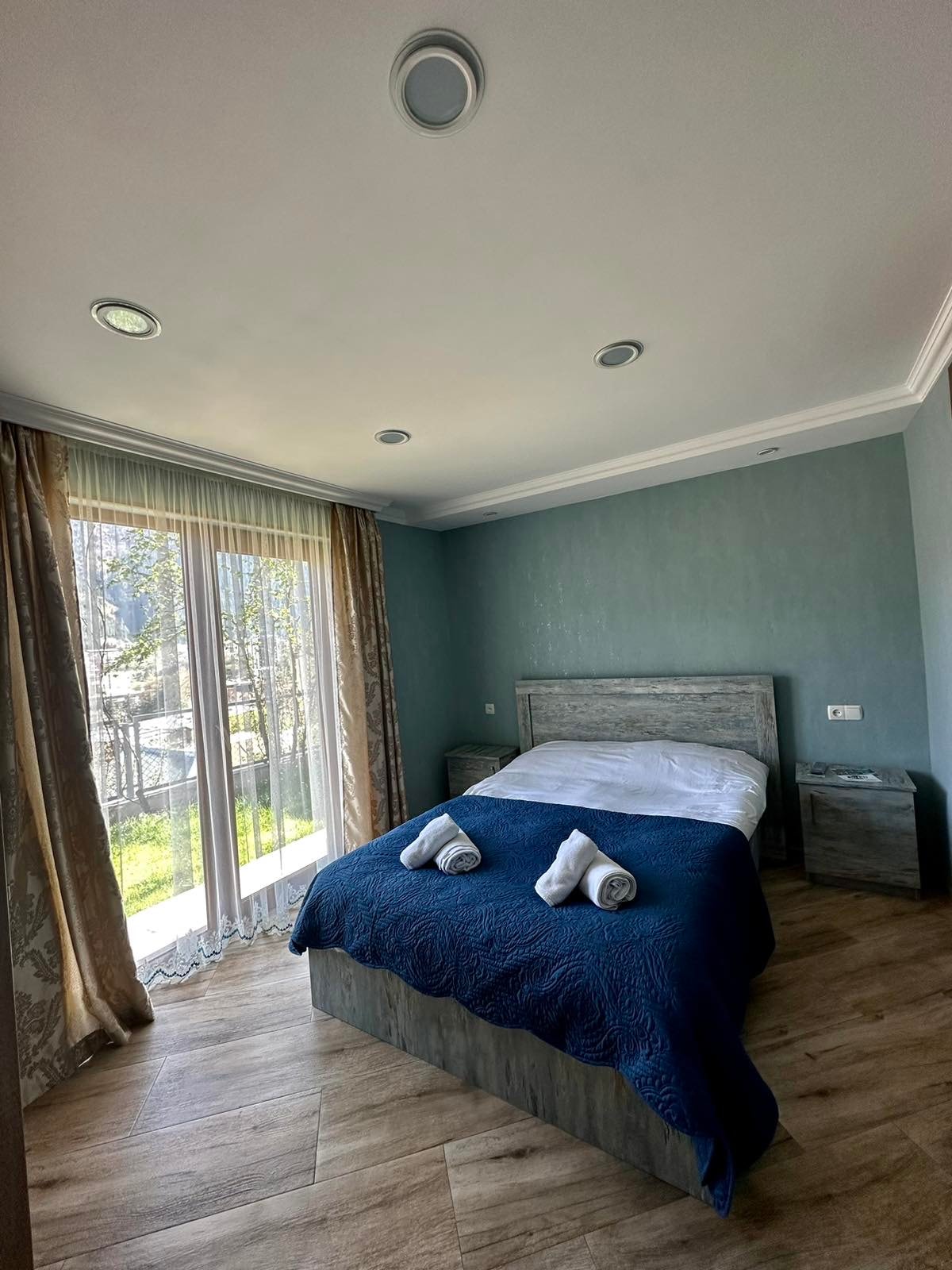 A light-filled bedroom features a spacious bed with a dark blue blanket. Large glass doors provide access to an outdoor area, while soft drapes frame the windows. Two towels are neatly placed on the bed, and a nightstand complements the contemporary design.