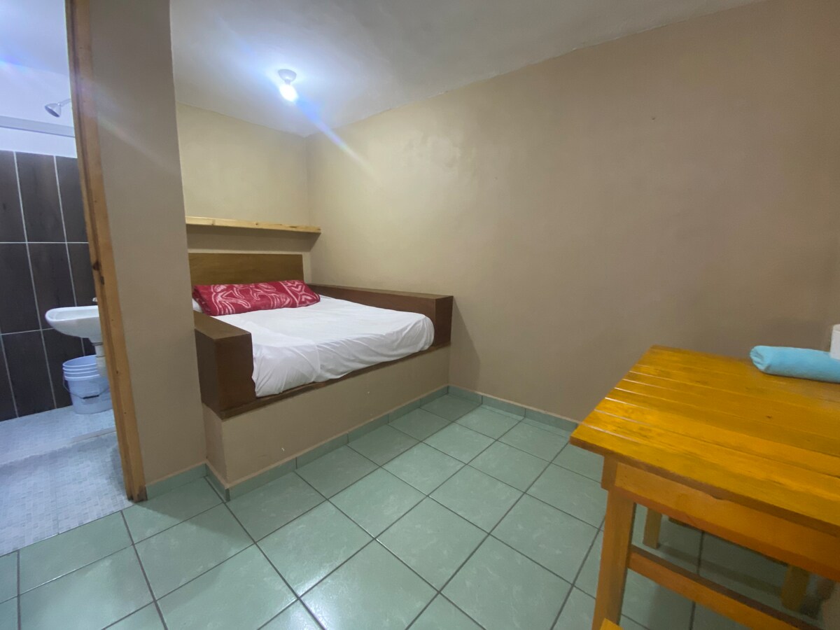 A sleeping area features a spacious bed with white linens and decorative pillows. A small wooden table is positioned nearby, and tiled flooring extends throughout the space. The open bathroom area is visible in the background, offering a functional design.