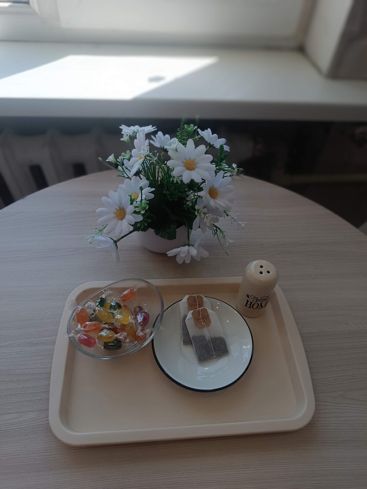 A light wooden table features a simple arrangement. A small bouquet of white daisies in a pot sits at the center. Nearby, a bowl of colorful candies and a plate holding a chocolate treat are positioned beside a salt shaker.