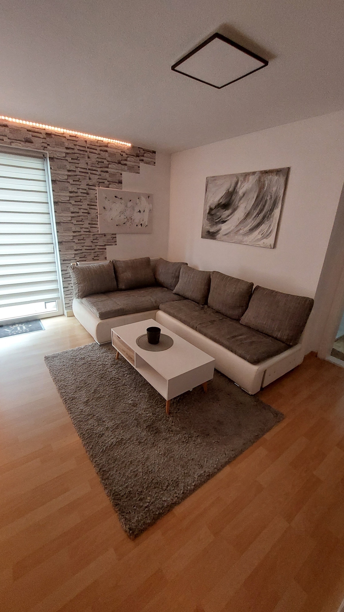 A comfortable living area is presented with a sectional sofa in muted tones. A light-colored coffee table sits on a soft area rug, complemented by a decorative black candle. Natural light filters through sheer window coverings, creating a serene atmosphere.