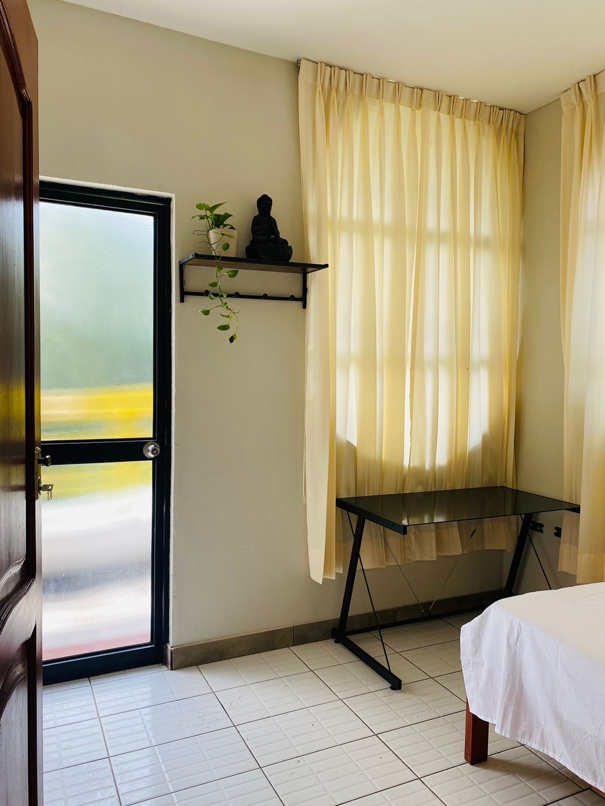 A bright and airy room features large windows with sheer curtains allowing natural light to fill the space. A small shelf holds a decorative statue and a plant, while a minimalistic glass desk is positioned against the wall, complementing the simple aesthetic.