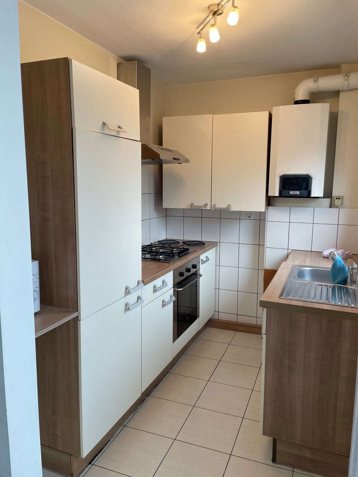The compact kitchen features sleek white cabinets with wooden accents, a gas stove, and a stainless steel sink. Simple tile backsplash complements the neutral color scheme, and soft overhead lighting illuminates the space, creating a functional cooking area.