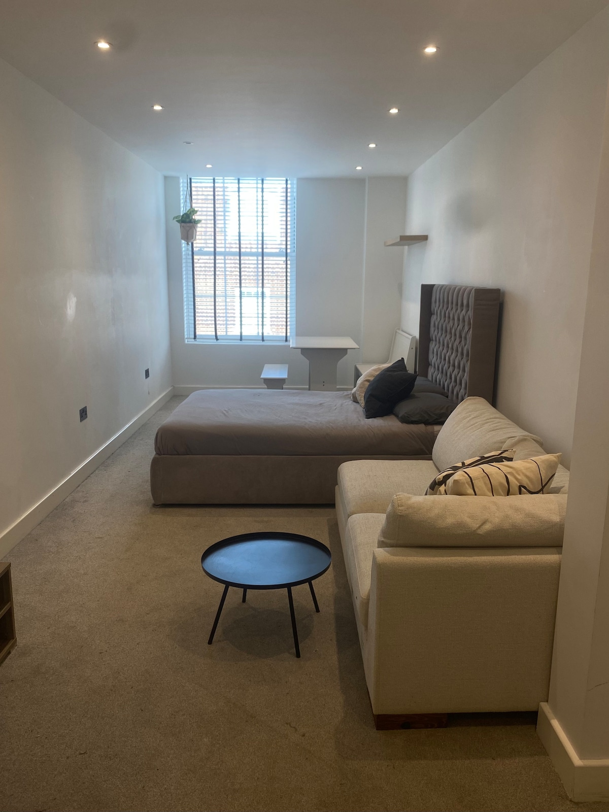 The spacious room features a layout with a double bed and a comfortable sofa. A small black table is positioned in the center, while a bright window with blinds allows natural light to fill the area. Soft carpeting enhances the inviting environment.