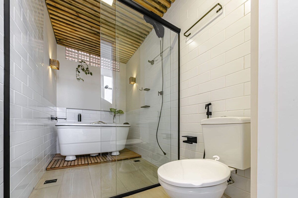 The bathroom features a modern glass shower enclosure alongside a freestanding bathtub. Natural light is accentuated by overhead wooden beams, and the space includes stylish lighting fixtures and greenery adding a touch of freshness. A toilet is positioned to the side.